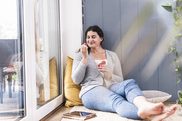 Junge Frau isst Erdbeere, während sie zu Hause am Fenster sitzt - UUF21943