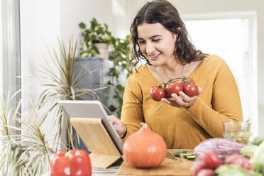 Smiling woman holding tomato while using digital tablet at home - UUF21947