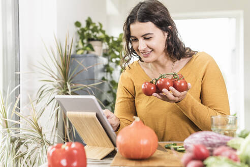 Lächelnde Frau, die eine Tomate in der Hand hält und ein digitales Tablet zu Hause benutzt - UUF21947