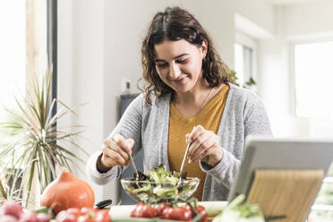 Lächelnde Frau bei der Zubereitung von Salat, während sie zu Hause am Tisch steht - UUF21950