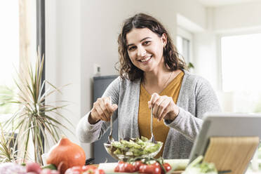 Smiling young woman making salad through digital tablet at home - UUF21952