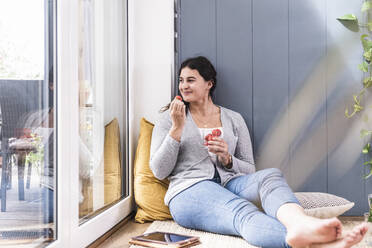 Lächelnde junge Frau isst Erdbeere, während sie zu Hause am Fenster sitzt - UUF21960