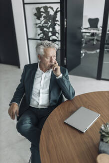Thoughtful businessman sitting on chair by table at office - GUSF04476