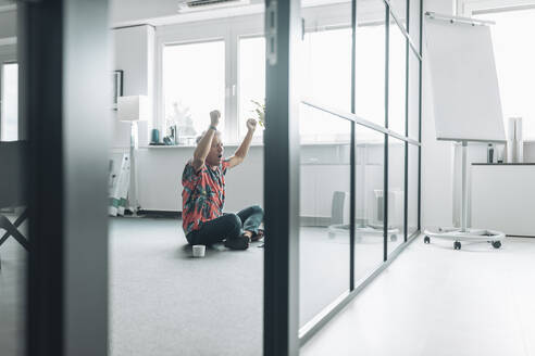 Businessman with arms raised sitting on floor at office - GUSF04488