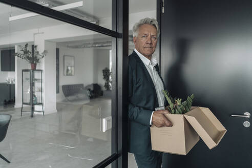 Businessman holding box of botany plant while leaning on glass wall at office - GUSF04528