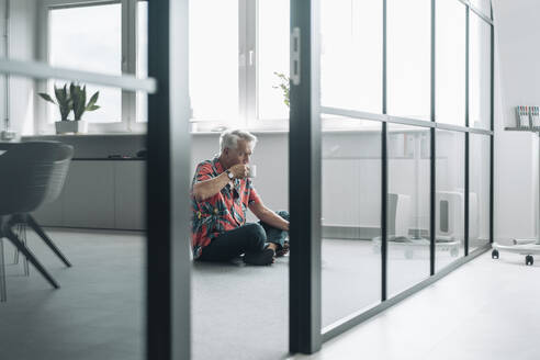 Businessman drinking coffee while sitting on floor at office - GUSF04536