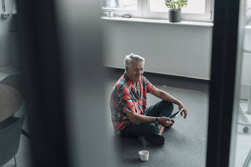 Smiling businessman using digital tablet while sitting on floor at office - GUSF04538