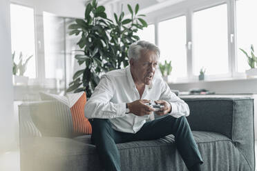 Älterer Geschäftsmann spielt konzentriert ein Videospiel, während er auf dem Sofa im Büro sitzt - GUSF04545