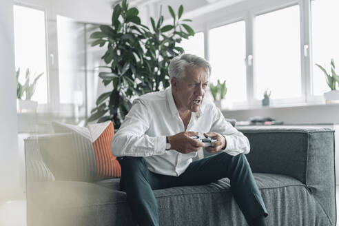Senior businessman playing video game with concentration while sitting on sofa at office - GUSF04545