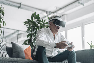 Senior businessman playing video game with virtual reality eyeglasses at office - GUSF04546