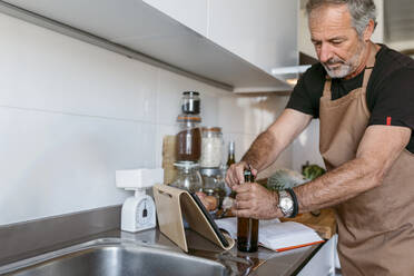 Mature man opening beer bottle while standing in kitchen - VABF03654