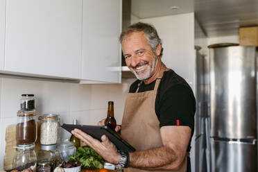 Lächelnder Mann, der eine Bierflasche und ein digitales Tablet hält, während er zu Hause in der Küche steht - VABF03655