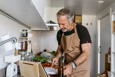 Mature man reading book while standing in kitchen at home - VABF03656