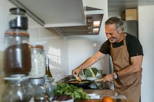 Glücklicher reifer Mann bereitet Nudeln zu, während er zu Hause in der Küche steht - VABF03660