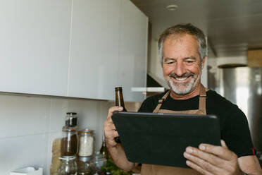 Lächelnder Mann, der eine Bierflasche hält, während er ein digitales Tablet benutzt, das in der Küche zu Hause steht - VABF03664