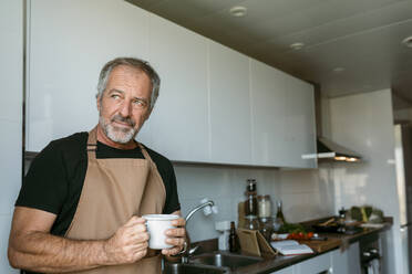 Reifer Mann, der wegschaut und eine Kaffeetasse hält, steht in der Küche zu Hause - VABF03666