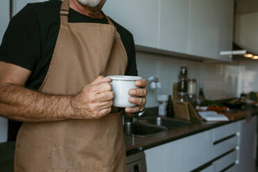 Mittelteil eines Mannes, der eine Kaffeetasse hält, stehend in der Küche zu Hause - VABF03667