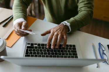 Mittelteil eines Mannes, der eine Kreditkarte benutzt, während er zu Hause am Laptop arbeitet - VABF03673