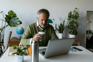 Älterer Mann trinkt Kaffee, während er zu Hause am Laptop arbeitet - VABF03674
