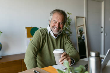 Smiling mature man holding disposable coffee cup sitting at home - VABF03678