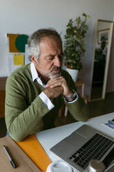 Mann mit Hand am Kinn, der einen Laptop benutzt, während er zu Hause sitzt - VABF03680