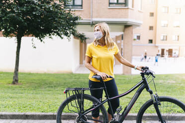 Mature woman wearing protective face mask looking away standing with bicycle on footpath in city - DGOF01561