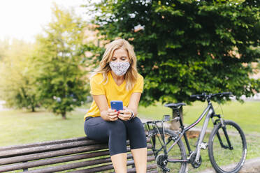 Woman wearing protective face mask using smart phone sitting on bench in city - DGOF01566
