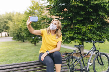 Woman taking selfie wearing protective face mask sitting on bench in city - DGOF01567