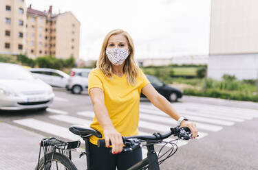 Frau mit Gesichtsschutzmaske steht mit Fahrrad auf der Straße in der Stadt - DGOF01572
