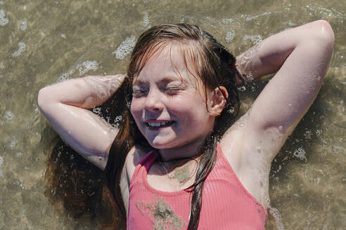 Girl lying on back with hands behind head in water during sunny day - OGF00603