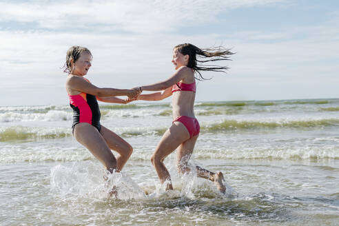 Friends holding hands enjoying while playing in water on sunny day - OGF00604