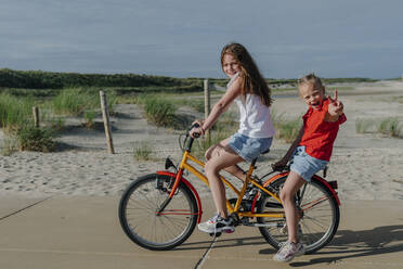 Mädchen nimmt jüngeres Geschwisterkind auf dem Fahrrad am Strand an einem sonnigen Tag - OGF00608