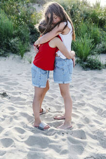 Sibling embracing while standing on sand at beach during sunny day - OGF00616