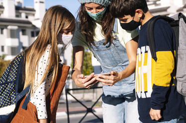 Mother and kids wearing protective face mask using smart phone standing in public park on sunny day - JCMF01549