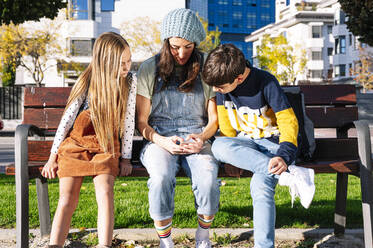 Mutter mit Kindern, die ein Smartphone benutzen, sitzt auf einer Bank in einem öffentlichen Park an einem sonnigen Tag - JCMF01553