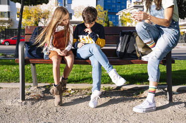 Niedriger Ausschnitt einer Mutter mit Kindern, die ihr Smartphone benutzen, während sie auf einer Bank in einem öffentlichen Park an einem sonnigen Tag sitzen - JCMF01556