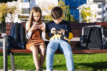 Bruder und Schwester benutzen ihr Smartphone, während sie auf einer Bank in einem öffentlichen Park bei Sonnenschein sitzen - JCMF01559