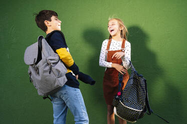 Brother and sister laughing while standing against green wall on sunny day - JCMF01562