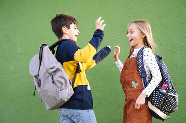 Brother and sister laughing doing high five while standing against green wall on sunny day - JCMF01563