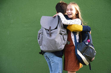 Brother and sister embracing each other while standing against green wall on sunny day - JCMF01564