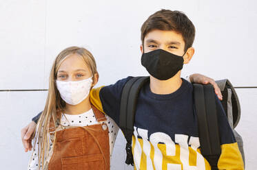 Brother and sister wearing protective face mask standing against wall on sunny day - JCMF01579