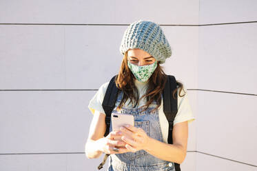 Mature woman using smart phone wearing protective face mask standing against wall on sunny day - JCMF01581