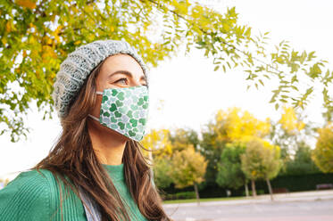 Mature woman looking away while wearing protective face mask standing in public park on sunny day - JCMF01584