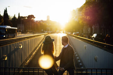 Couple looking away while sitting on bridge railing together at city during sunset - OCMF01797