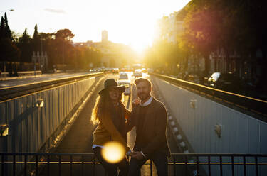 Couple holding hands while sitting on bridge railing together at city during sunset - OCMF01798