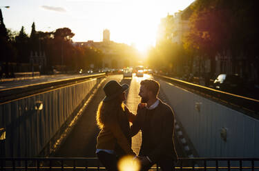 Couple looking at each other while sitting on bridge railing at city during sunset - OCMF01799