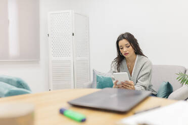 Junge Frau, die ein digitales Tablet benutzt, während sie auf der Couch im Büro sitzt - MRRF00616