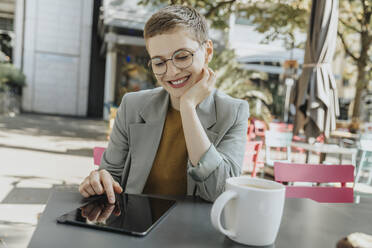 Frau mit digitalem Tablet sitzend mit Hand am Kinn in einem Straßencafé - MFF06710