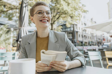 Smiling woman looking away while reading book sitting in sidewalk cafe - MFF06712