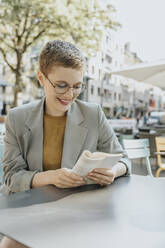 Lächelnde Frau, die in einem Straßencafé an einem sonnigen Tag ein Buch liest - MFF06713
